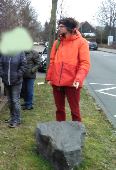 Der stellvertretende Fraktionssprecher der GRÜNEN, Benjamin Beckmann, deutet mit seiner Hand auf einen Felsbrocken, der auf einer straßenbegleitenden Grünfläche liegt.