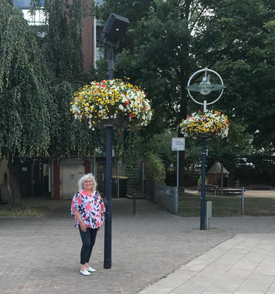 Frau Blotenberg steht auf dem Aplerbecker Marktplatz vor einer Blumenampel. Im Hintergrund ist eine weitere Blumenampel zu sehen.